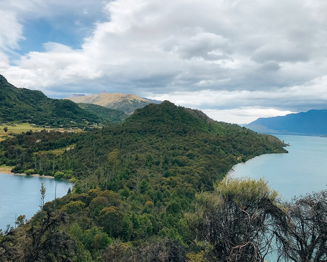 A view of a lush, green hill surrounded by a lake, with mountains in the background under a cloudy sky.