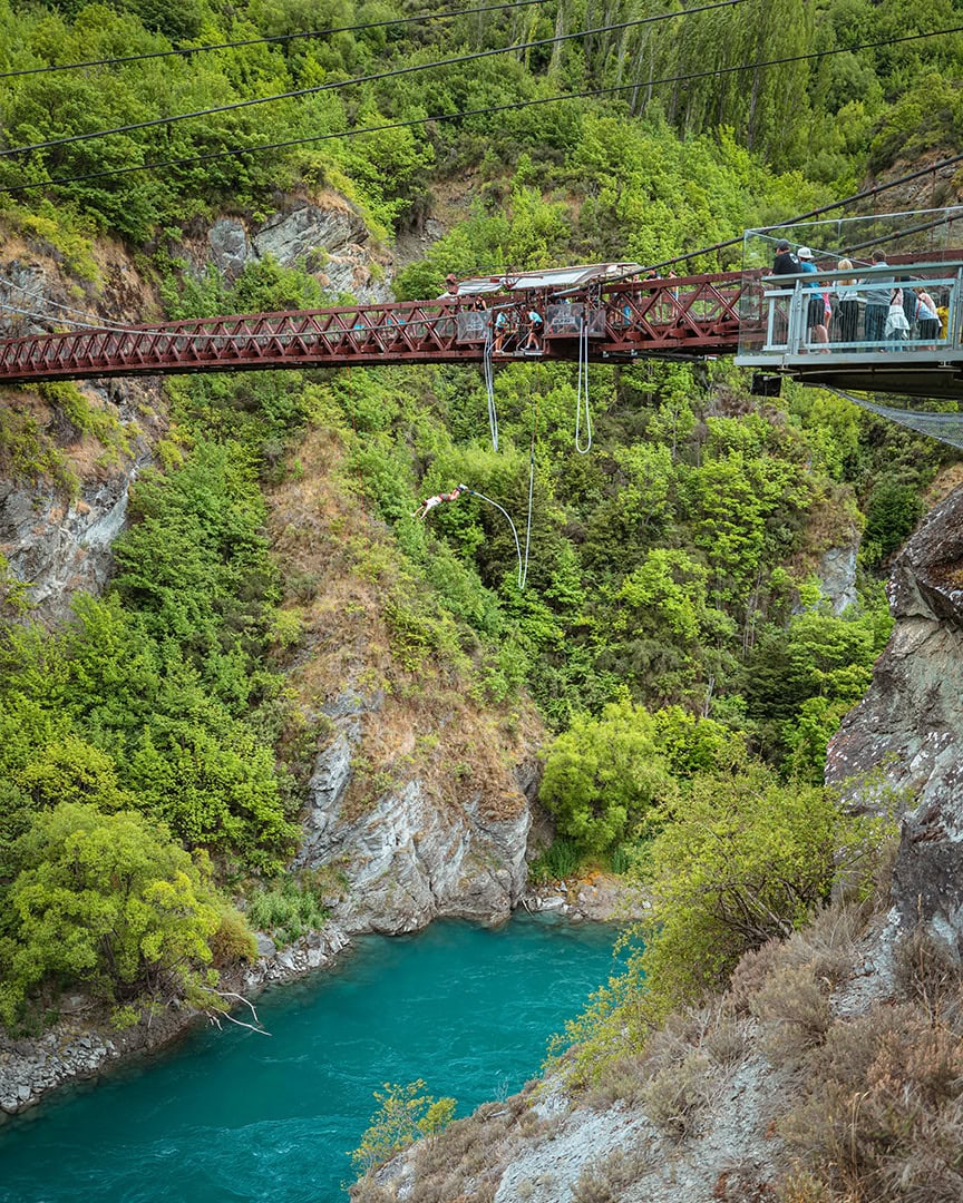 A person mid-air bungee jumping off a bridge over a turquoise river with green hillsides. Kawarau Bridge Bungy Queenstown.
