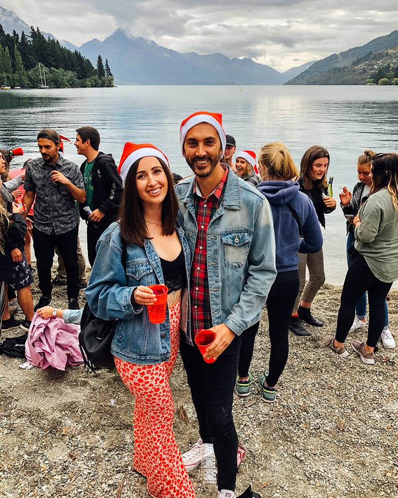 A couple wearing Santa hats holding red cups, surrounded by other people near a lake with mountains in the background. Queenstown at Christmas.