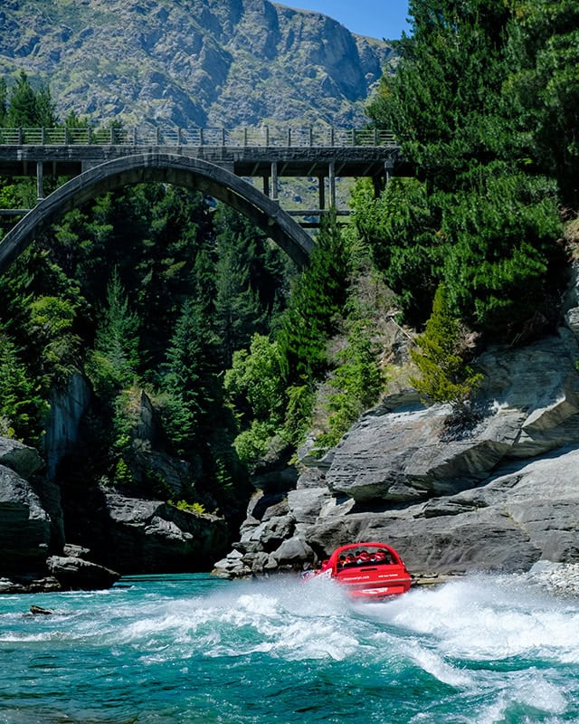 A red jet boat speeding through a narrow river gorge with a stone bridge above and dense pine trees around.