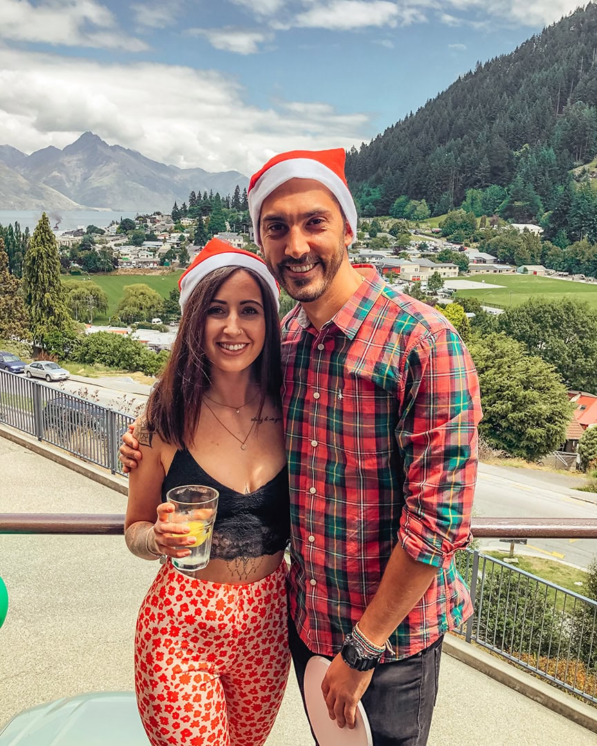 A smiling couple in Santa hats with a lake, mountains, and green forest as the background. Christmas in Queenstown.