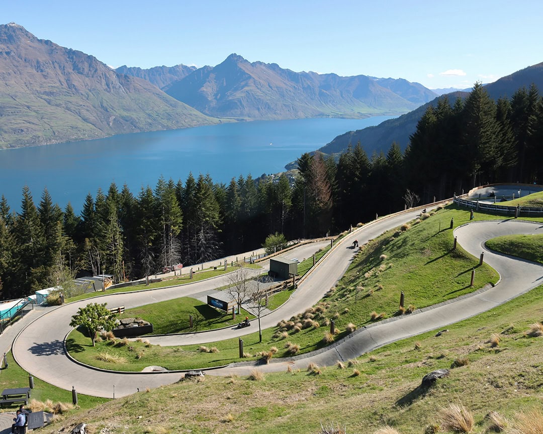 A winding luge track on a green hillside with a lake and mountain view in the background.