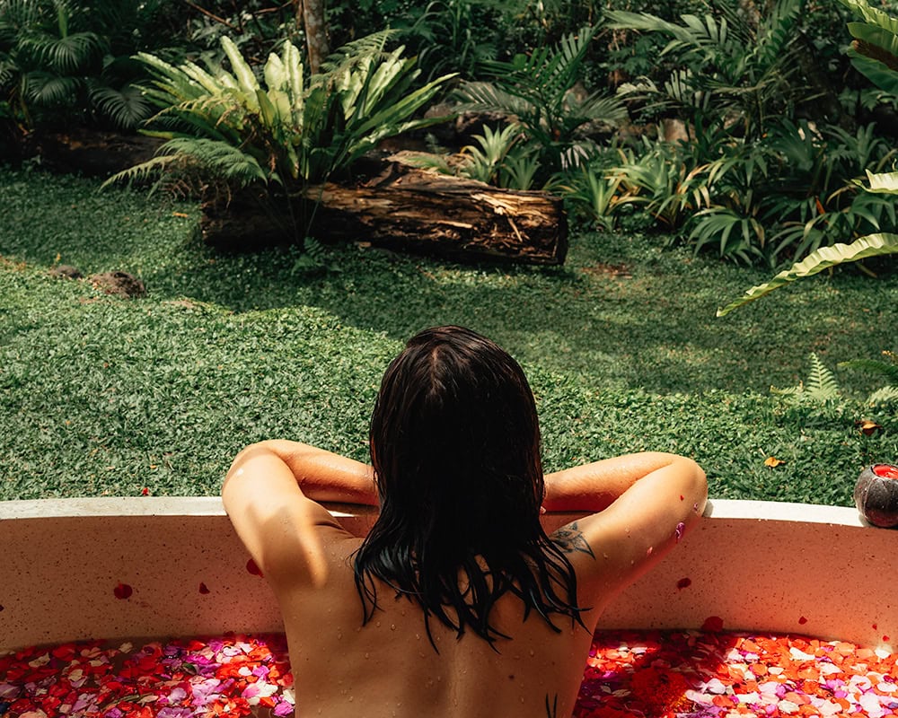 A woman relaxes in an outdoor stone bath filled with pink and red flower petals, gazing out at lush green plants and tropical trees.
