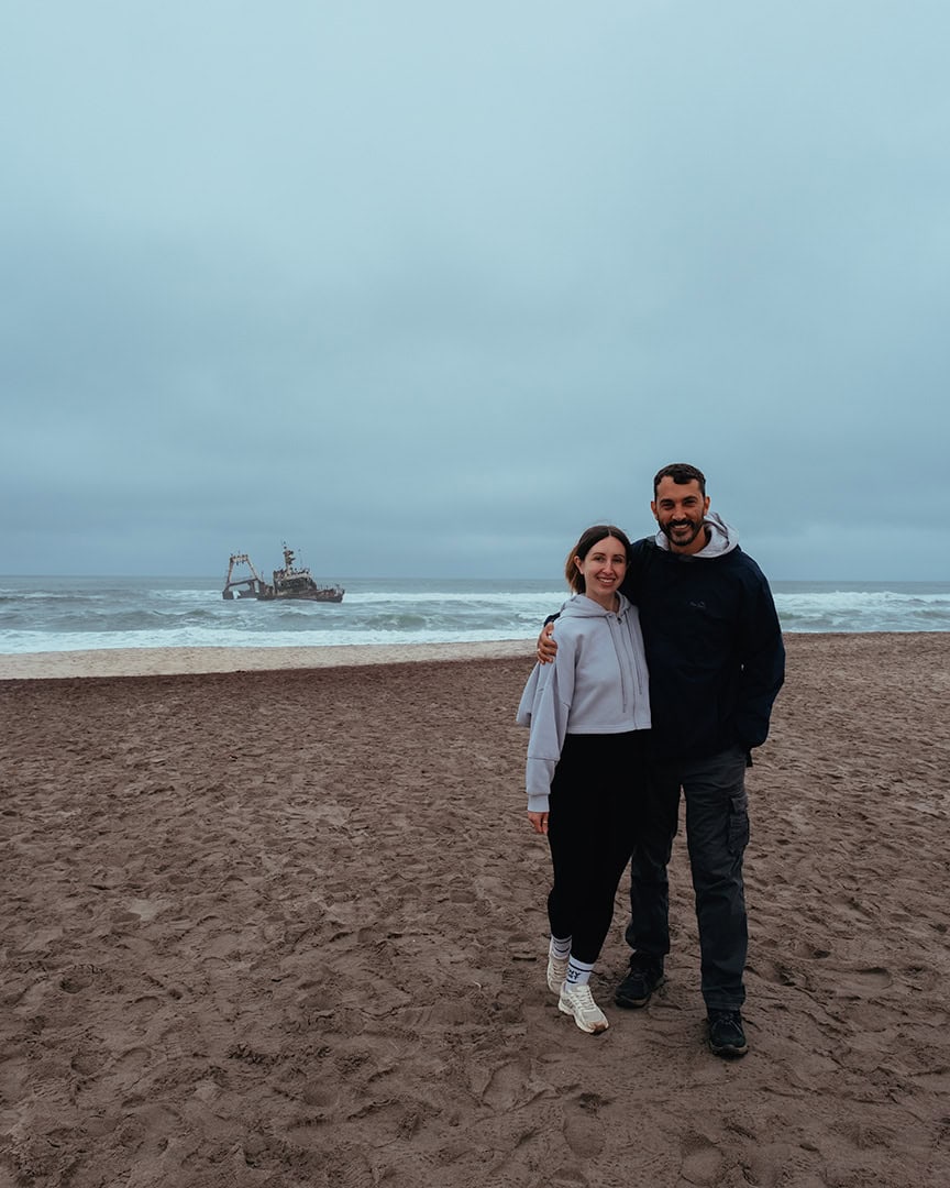 Couple standing on a sandy beach with waves crashing behind them. A shipwreck is visible in the ocean in the distance.