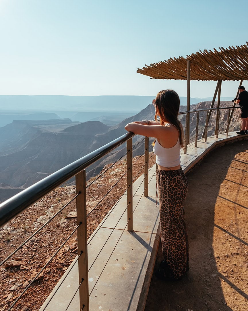 Woman leaning on a railing at a canyon viewpoint overlooking a vast rocky valley. A wooden shade structure stands above the viewing platform.