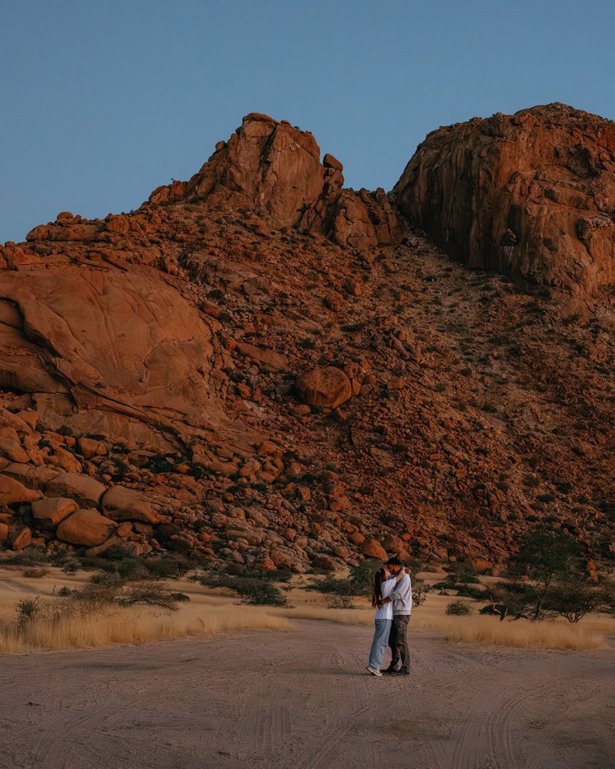 Couple standing and hugging in a sandy desert with towering red rock cliffs in the background.