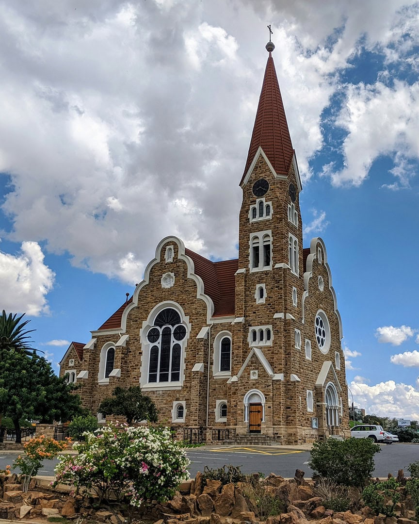 Historic stone church with a tall red roof steeple and arched windows under a partly cloudy sky. Flowers and rocks sit in the foreground near the street.