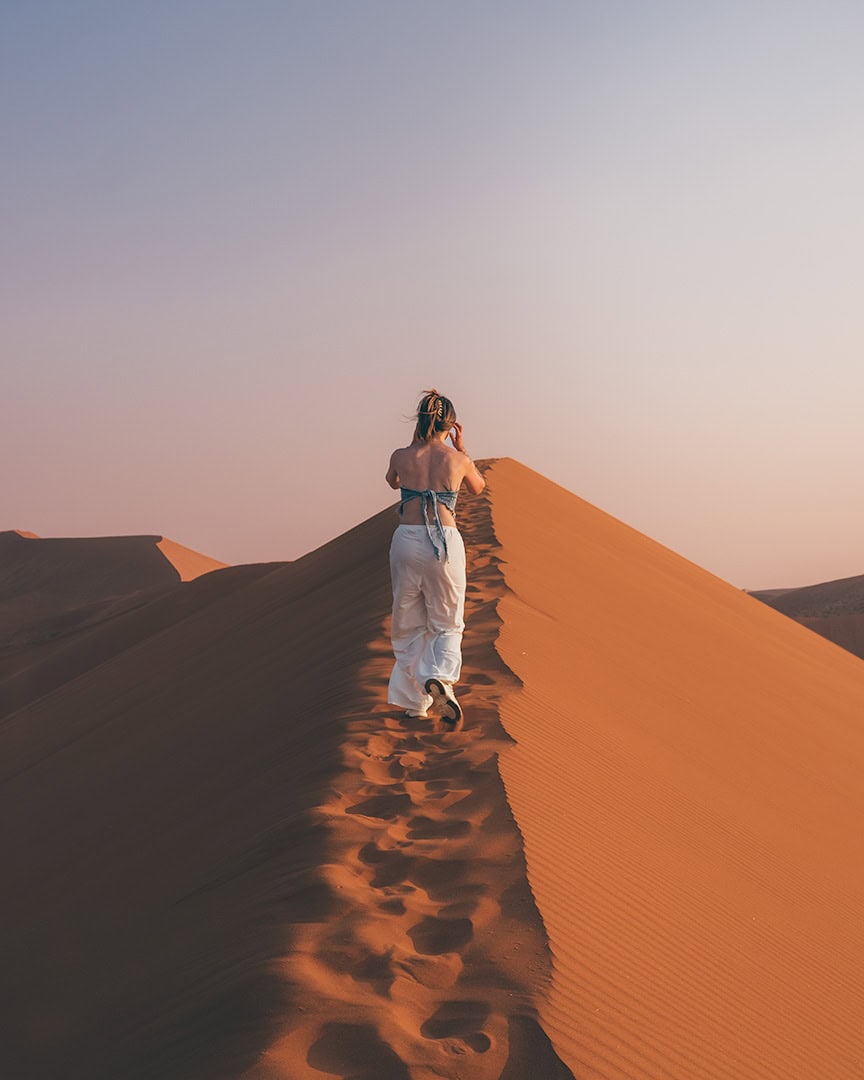 Woman walking along the ridge of a tall orange sand dune with footprints trailing behind her. The sky is clear and the desert stretches into the distance.