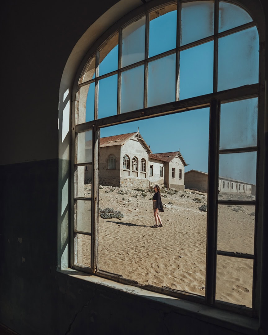 View through an arched window of a sandy desert scene with old buildings outside. A woman walks across the sand in front of the structures.