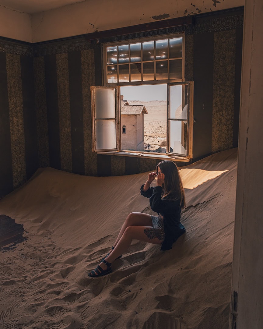 Woman sitting inside an abandoned room filled with sand dunes blowing in through an open window. Sunlight streams in and reveals the desert outside.