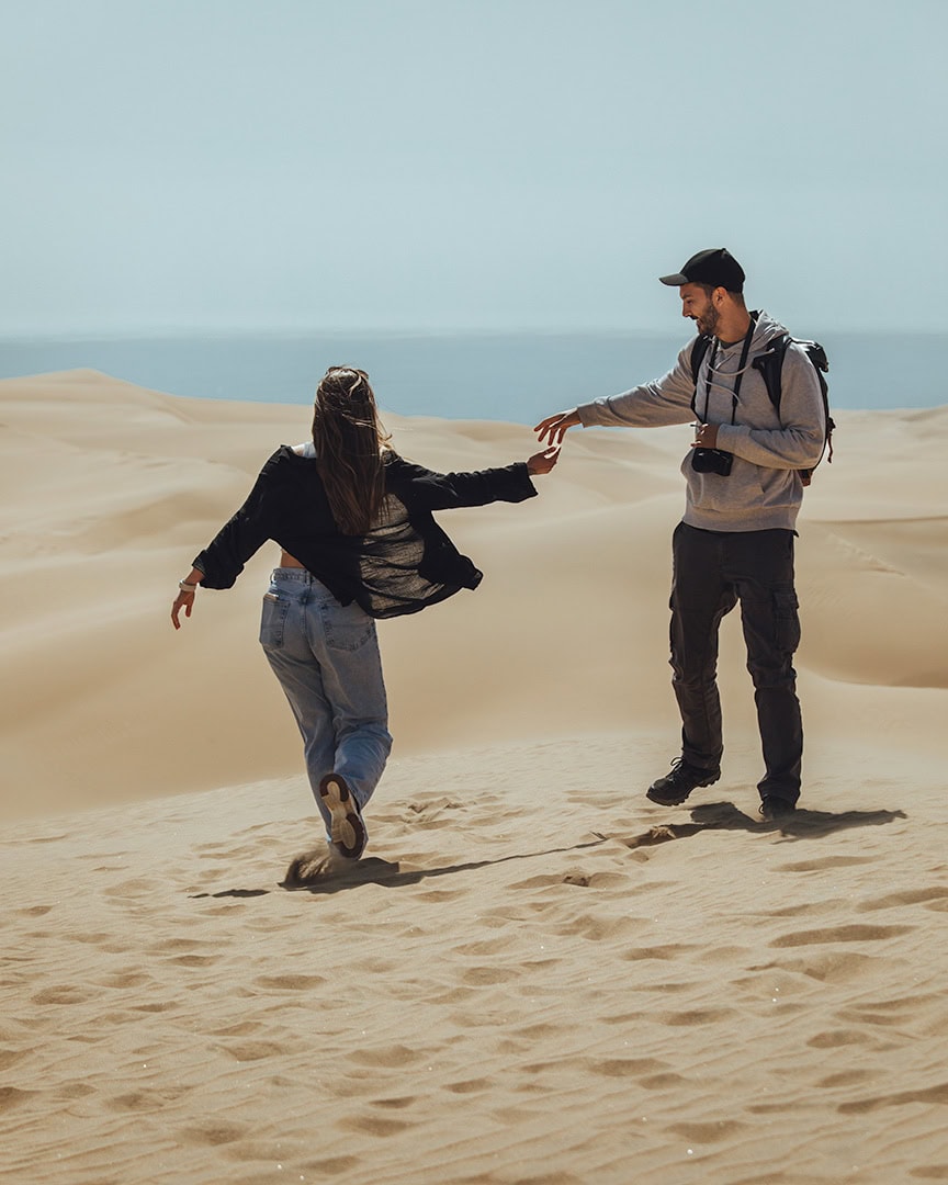 Couple walking hand in hand on sand dunes in the desert. Large rolling dunes stretch behind them under a clear blue sky.