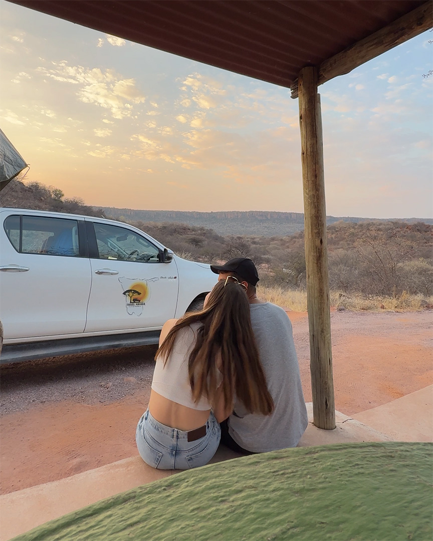 Couple sitting close together on a porch at sunset overlooking a dry savanna landscape. A white safari vehicle is parked nearby.