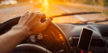 Driver's hand on steering wheel with phone and dashboard in sunlight.