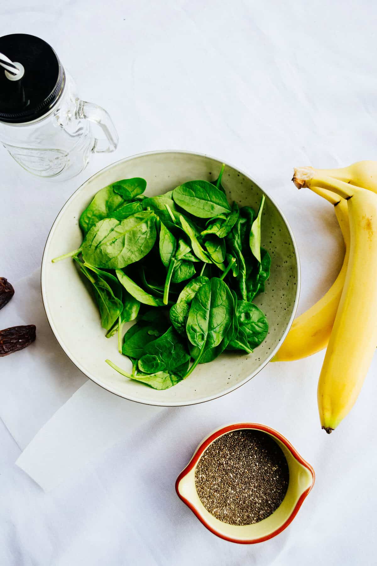 Fresh spinach in a white bowl with bananas, chia seeds, and a water bottle on a white table ready for healthy eating.