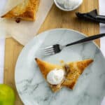 Warm apple pie slice with golden crust topped with vanilla ice cream on a marble plate, served with additional ice cream and fresh green apples on a wooden cutting board, perfect for a sweet dessert.