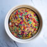 Colorful sprinkled birthday cake in a white bowl on a marble surface.