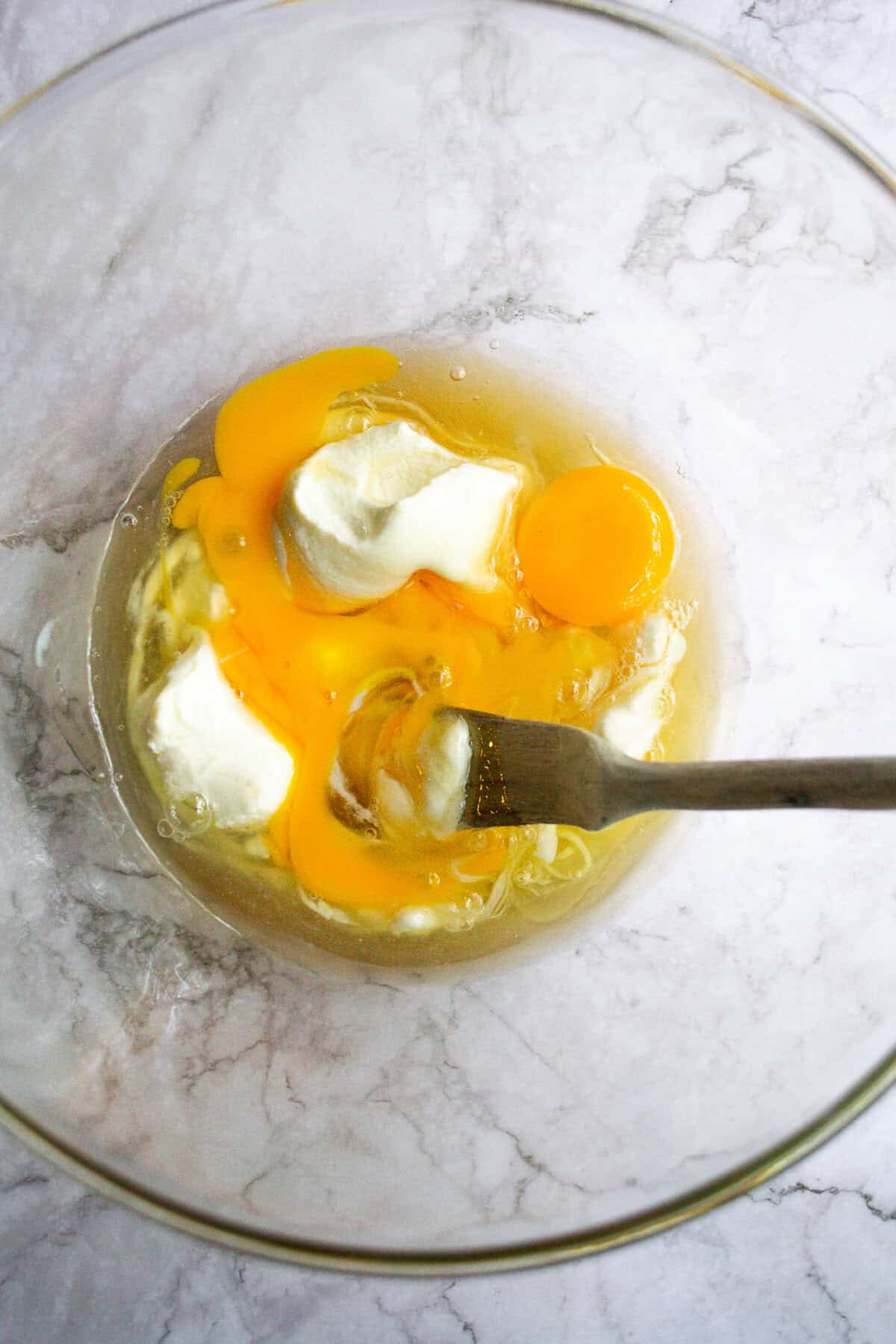 Creamy scrambled eggs being mixed in a glass bowl with a fork, showcasing fresh eggs and dairy ingredients for a simple breakfast recipe.