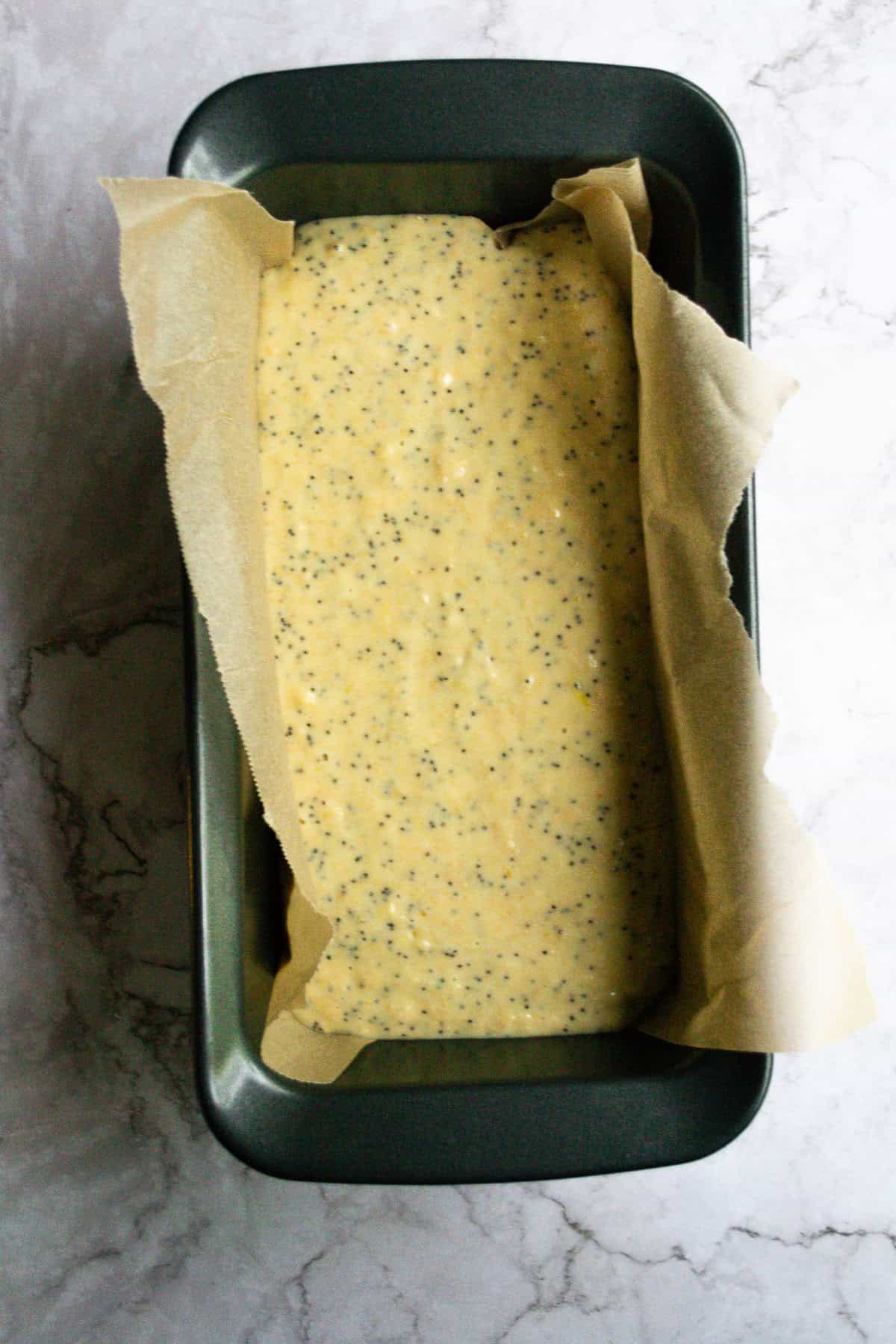 Creamy yellow custard batter with poppy seeds in a baking pan lined with parchment paper, ready to be baked for a delicious homemade treat.