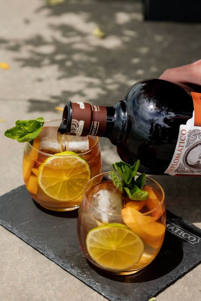 Refreshing Aperol Spritz cocktail with lemon, mint, and ice served in two glasses on a black slate board. Perfect for summer, featuring a bottle of Aperol labeled "DILOMATICO" being poured over ice.