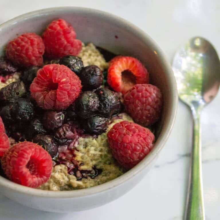 Fresh berry oatmeal bowl with raspberries and blueberries for healthy breakfast, served in a white bowl with a silver spoon on a marble surface, simple nutritious food photo.