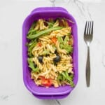 Creamy vegan pasta salad with fresh cherry tomatoes, black olives, and arugula in a purple container on a white marble surface. Perfect for healthy, quick lunch ideas.