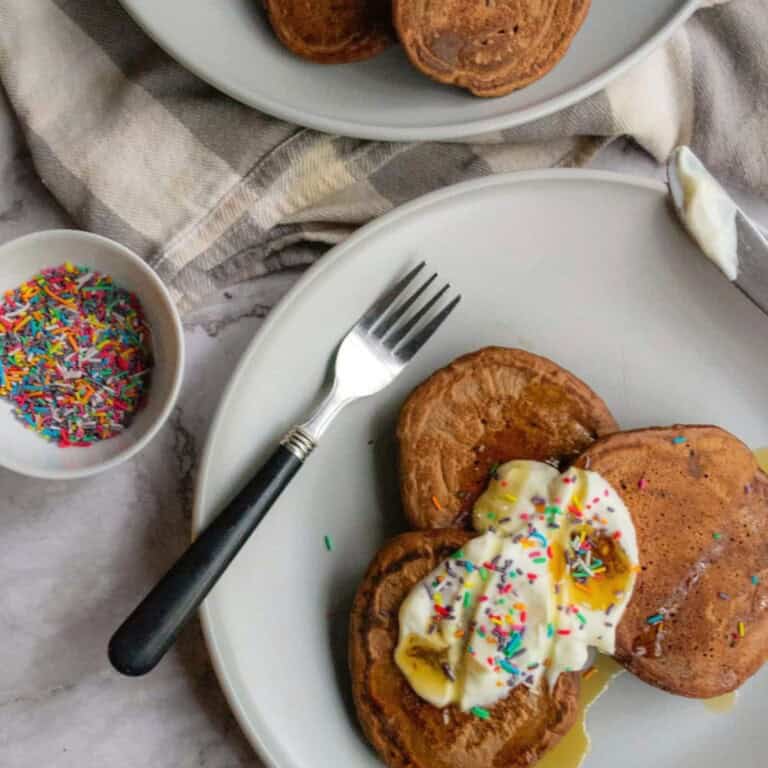 Fluffy pancakes with whipped cream and colorful sprinkles served on white plates, cozy table setting with a fork, bowl of sprinkles, and drizzled syrup, perfect for homemade breakfast or brunch recipes.