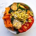 Fresh vegetable salad with roasted zucchini, cherry tomatoes, shredded carrots, sunflower seeds, and edamame, served in a white bowl on a marble surface. Perfect for healthy eating and vegan meal ideas.