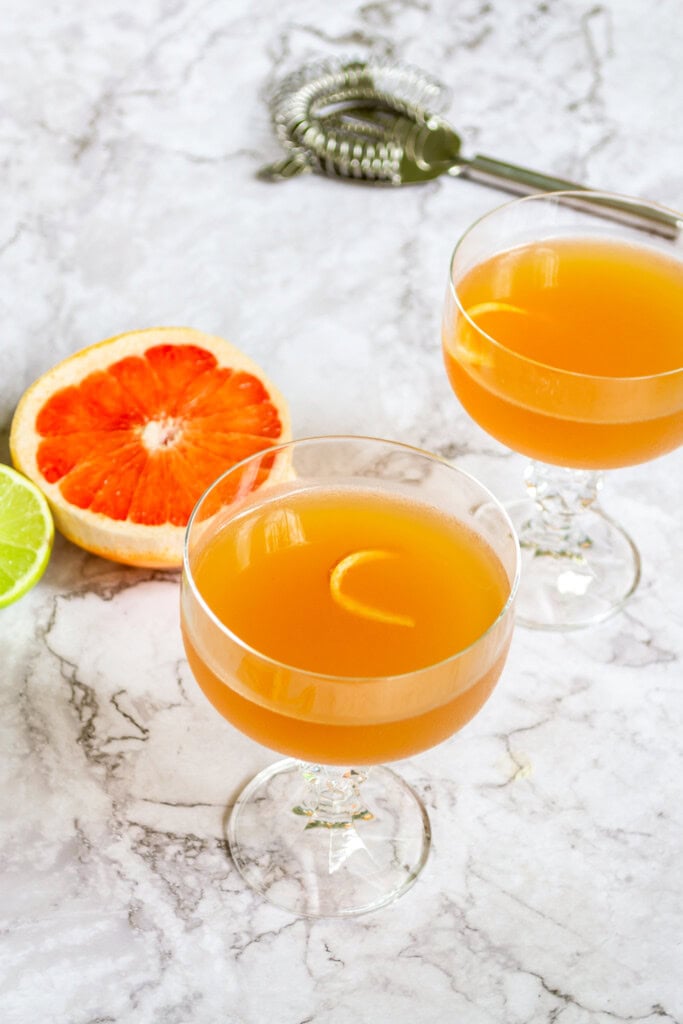 Two grapefruit daiquiris on a white marble table with a grapefruit and cocktail strainer in the background.
