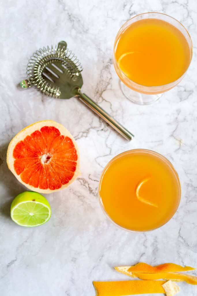 Top view of grapefruit daiquiri ingredients and cocktails. There is a lime, grapefruit, cocktail strainer, and grapefruit peel on the table with the two cocktails.