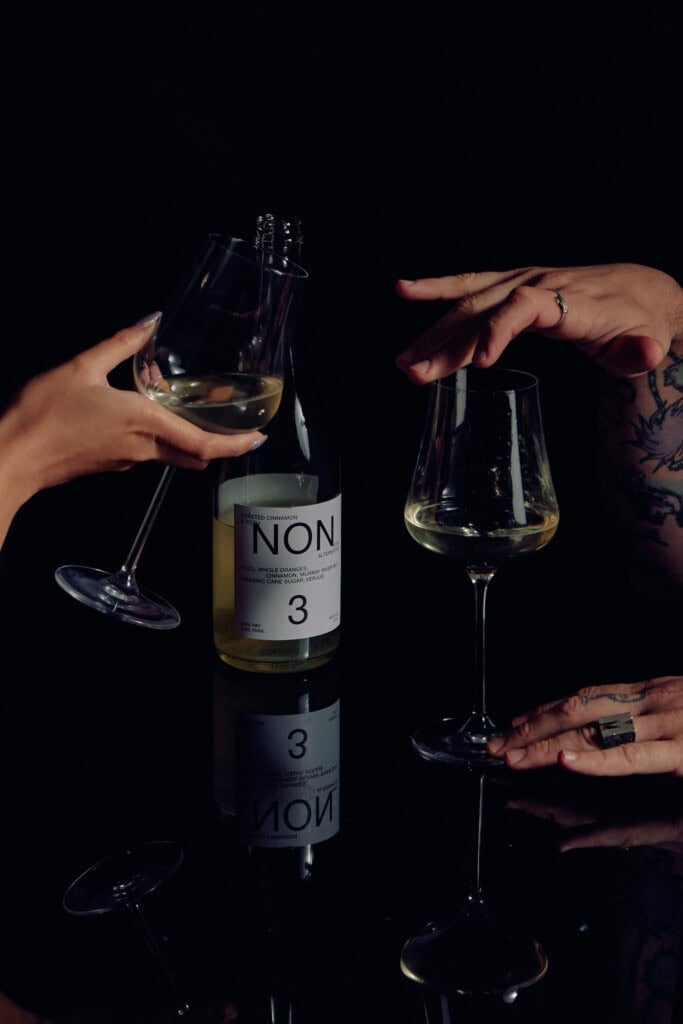 Bottle of NON alternative spirit with two glasses of clear liquid, hands reaching for the glasses, black background, modern and minimalist aesthetic, popular for cocktails and social drinking.