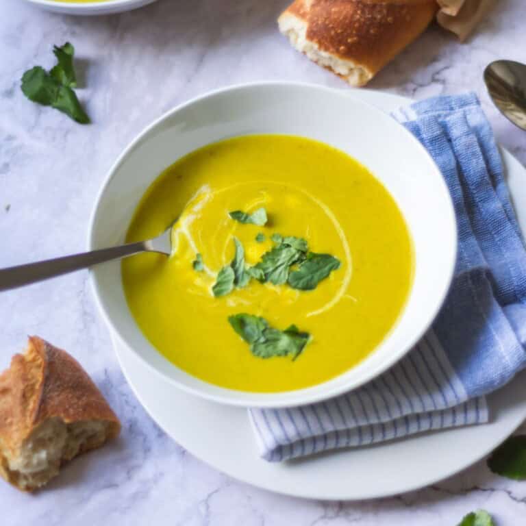 Creamy yellow vegetable soup garnished with fresh basil leaves, served in a white bowl, with crispy bread on the side, perfect for a healthy meal from Simply Fresh Foodie.