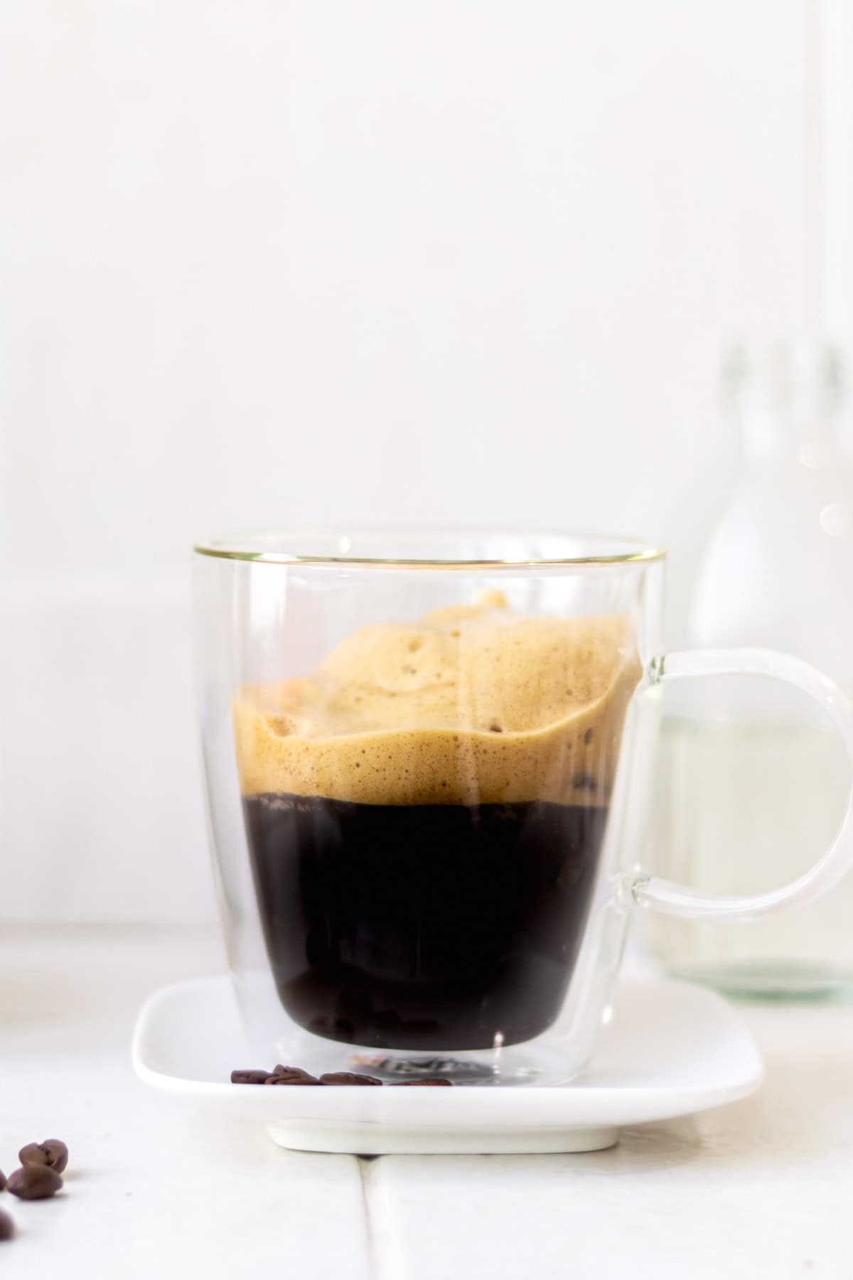 Rich black coffee in a transparent glass mug with foam on top, on white surface with a few coffee beans scattered, minimalist style.