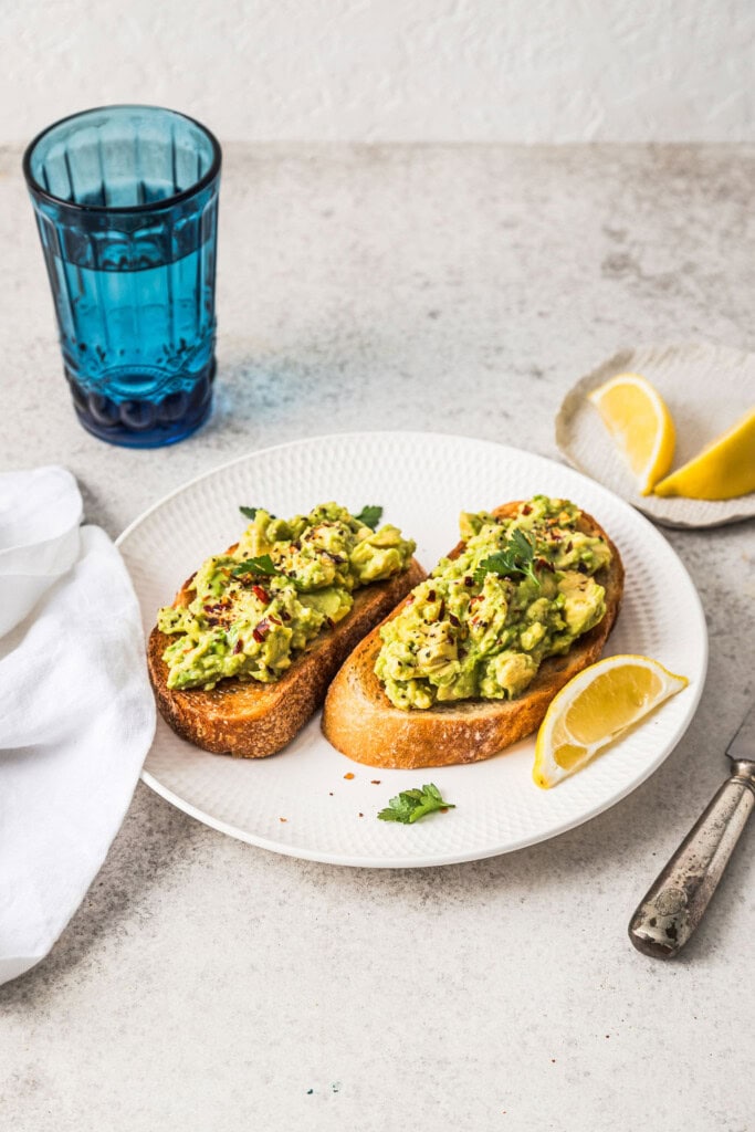 Front angle shot of avocado toast on a white plate, with lemon slices and a glass of water in the background.