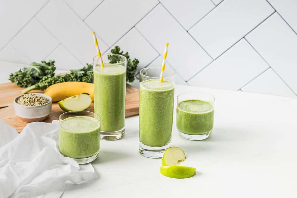Landscape photo of two kale apple smoothies on a white table, with the ingredients in the background on a wooden cutting board including kale, hemp seeds, apple and banana.