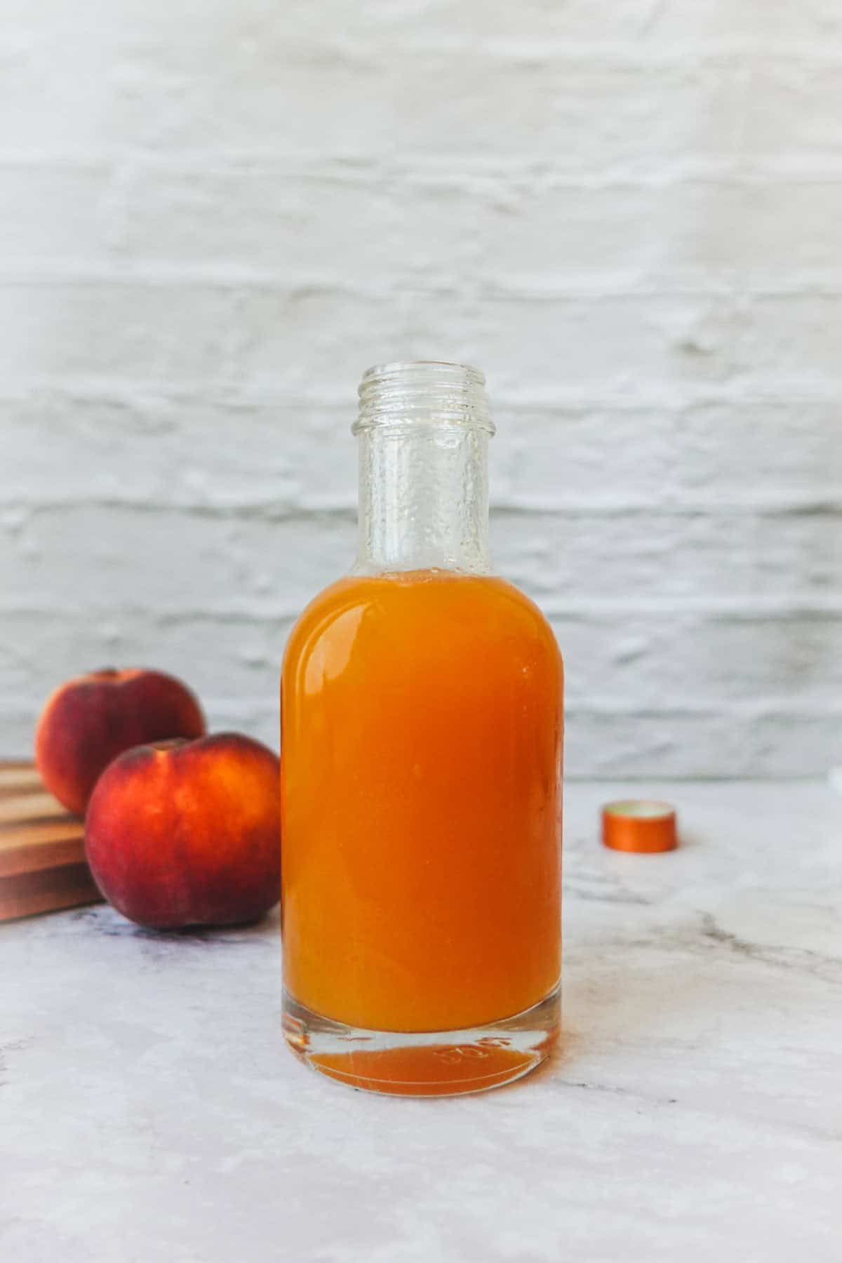 Straight-on shot of peach syrup in a glass bottle with peaches in the background.
