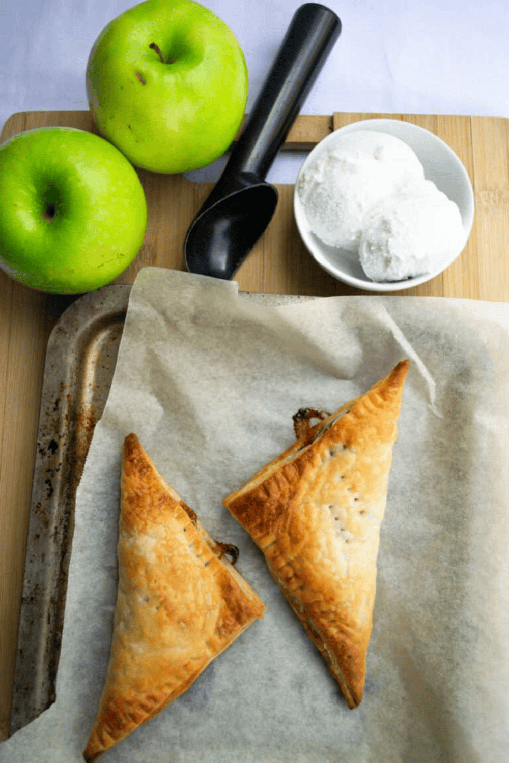 Overhead shot of two vegan apple turnovers on a baking tray with Granny Smith apples and a scoop of ice cream pictured above.