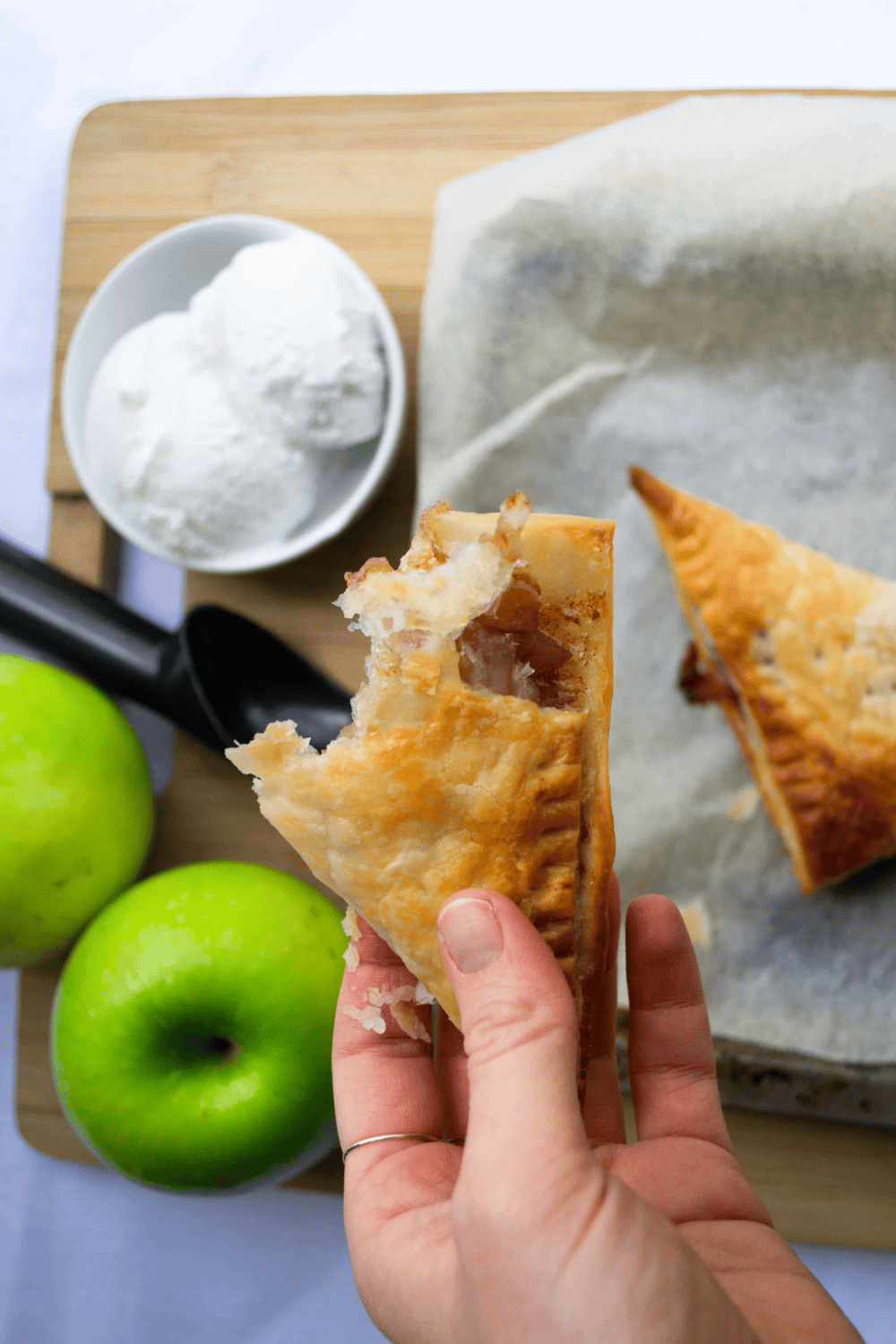 Hand holding half of a vegan apple turnover with a bite out of it with apples and a scoop of vanilla ice cream in the background.