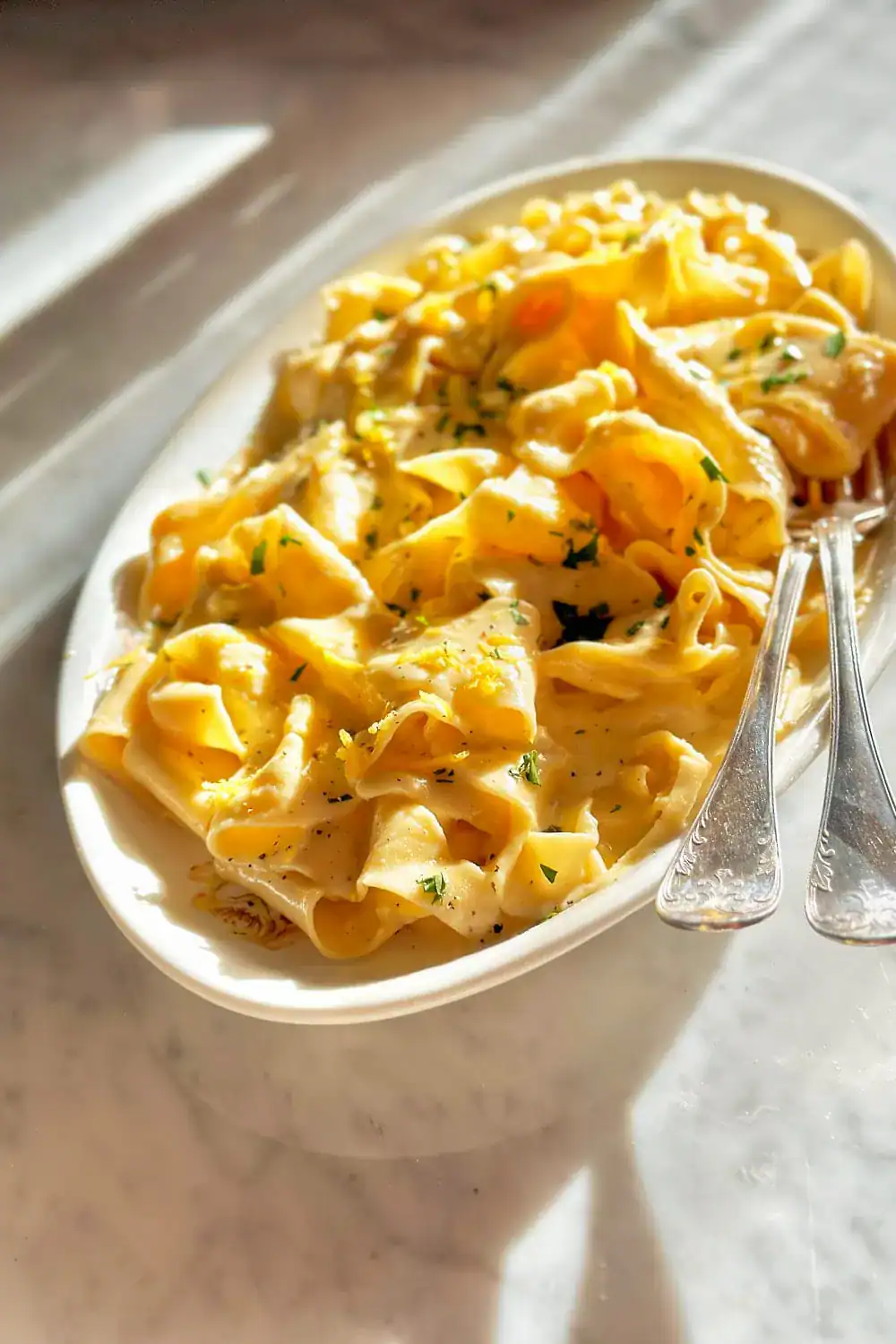 Creamy fettuccine alfredo pasta with herbs in a white bowl, served on a marble table with vintage silverware, highlighting delicious homemade comfort food.