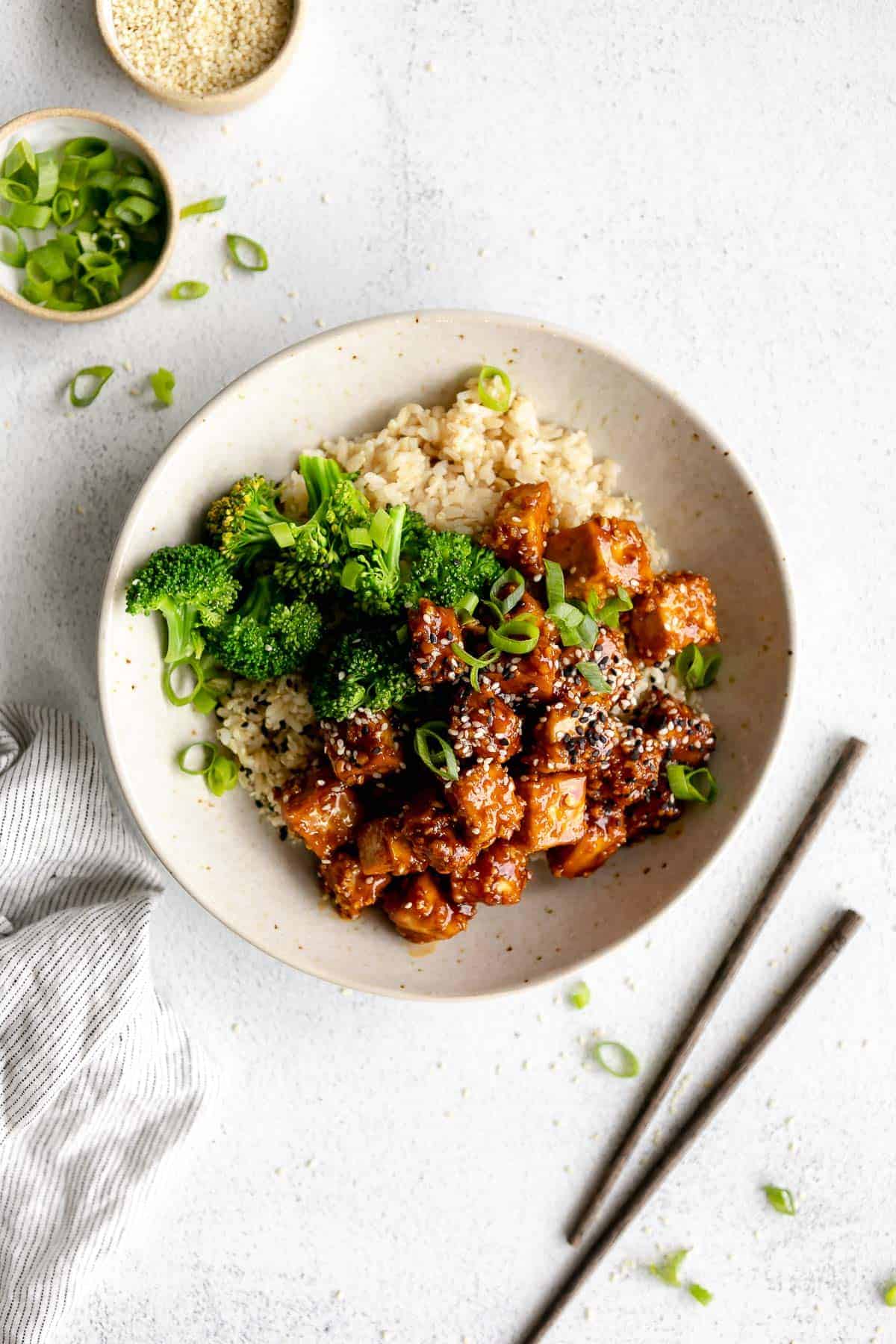 Overhead view of a sesame tofu bowl with brown rice and broccoli, in a white bowl on a white table.