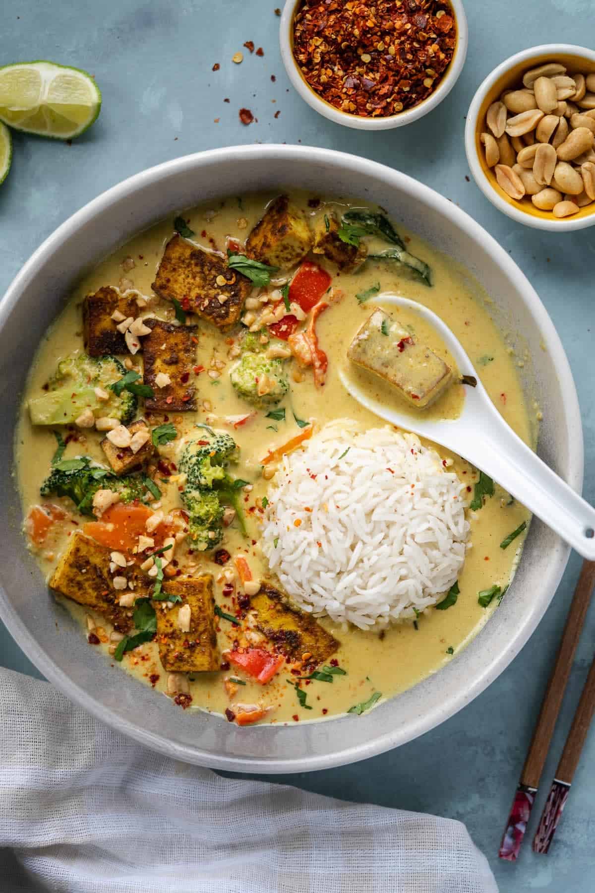 Savory Thai green curry with tofu, vegetables, and jasmine rice served in a white bowl, garnished with chopped nuts and fresh herbs, with lime wedges and chili flakes on the side.