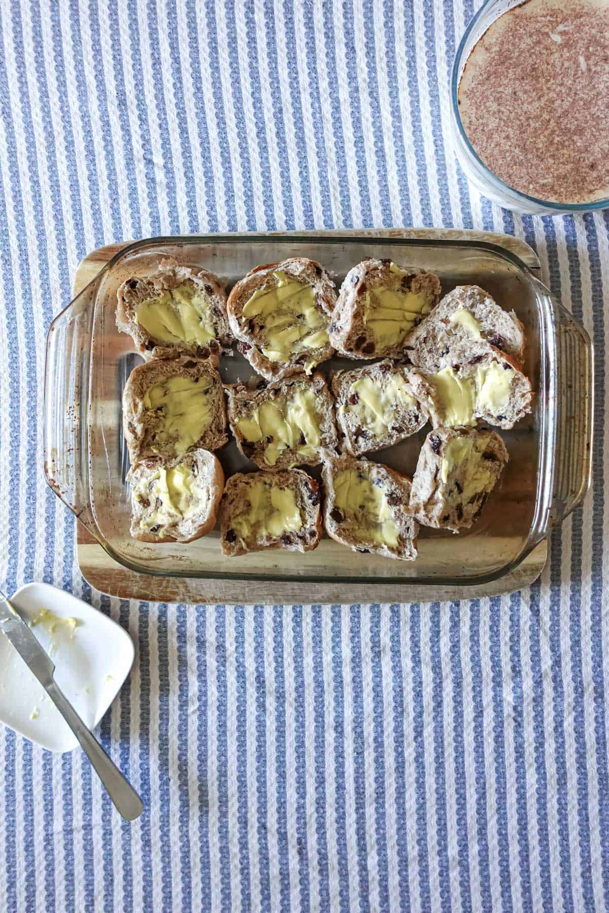 Butter stained bread slices filled with thick butter spreads on a glass baking dish with a blue striped cloth background and a small white bowl with a butter knife for easy spreading.