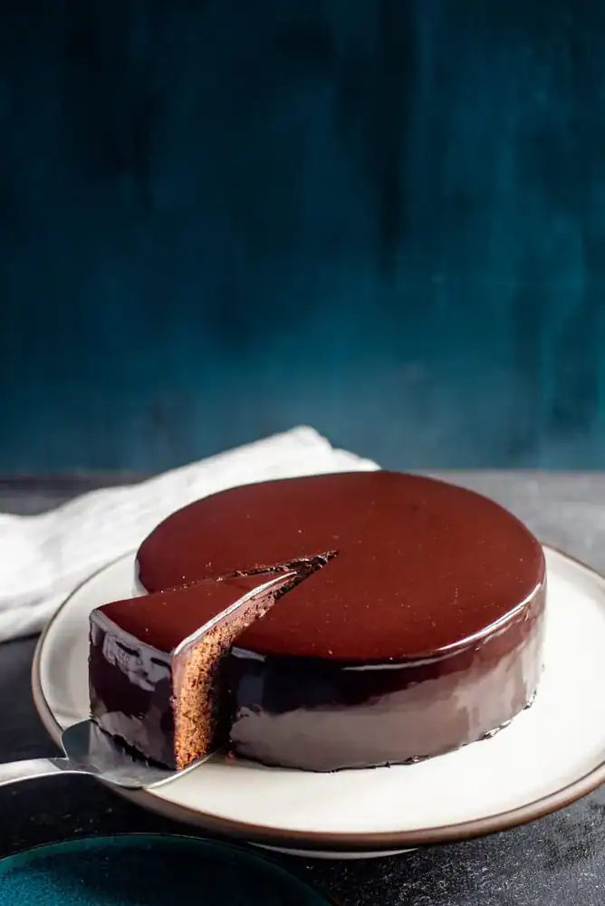 Close up of a cake with chocolate mirror glaze sitting on a white plate, with a slice being cut out of it.