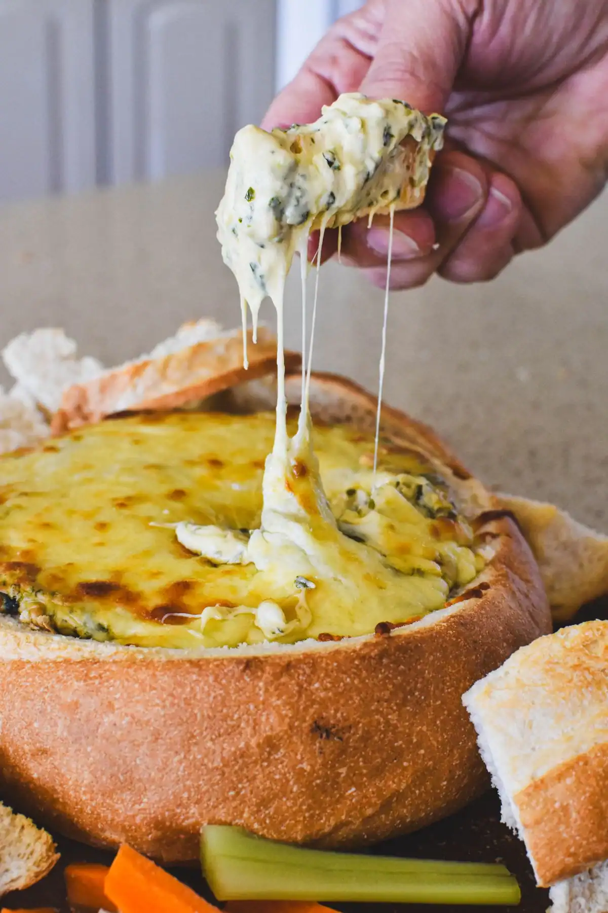 Creamy spinach dip served in a bread bowl, cob loaf-style with melted cheese and fresh vegetable sticks.