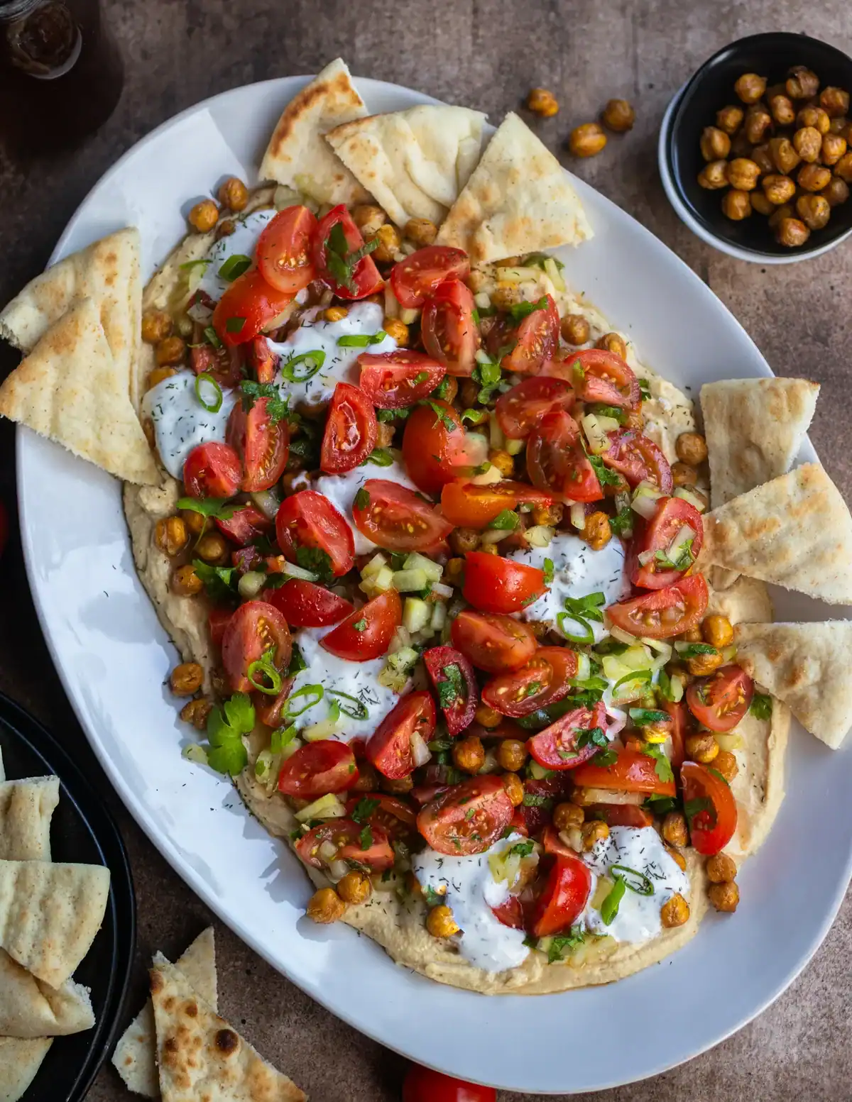 Fresh vegetarian hummus board platter topped with cherry tomatoes, chickpeas, yogurt sauce, and chopped scallions.