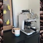 A white espresso machine sitting on top of a black cabinet, with a cup of coffee in the foreground and coffee-themed poster in the background.