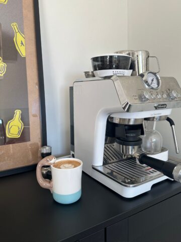 A white espresso machine sitting on top of a black cabinet, with a cup of coffee in the foreground and coffee-themed poster in the background.