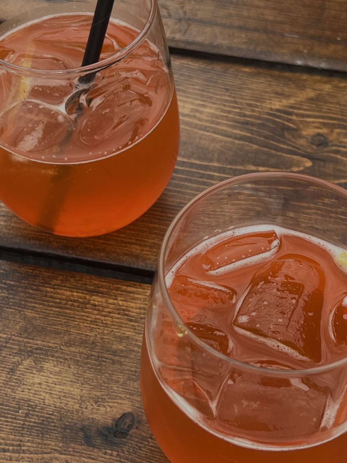 Close up of two glasses of red-orange coloured kombucha with ice on a wood table.