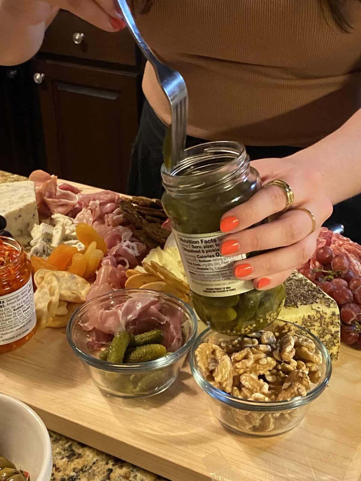 Food photography of a woman spooning pickles out of a jar onto a savoury charcuterie board with fruit, pickles and cheeses.