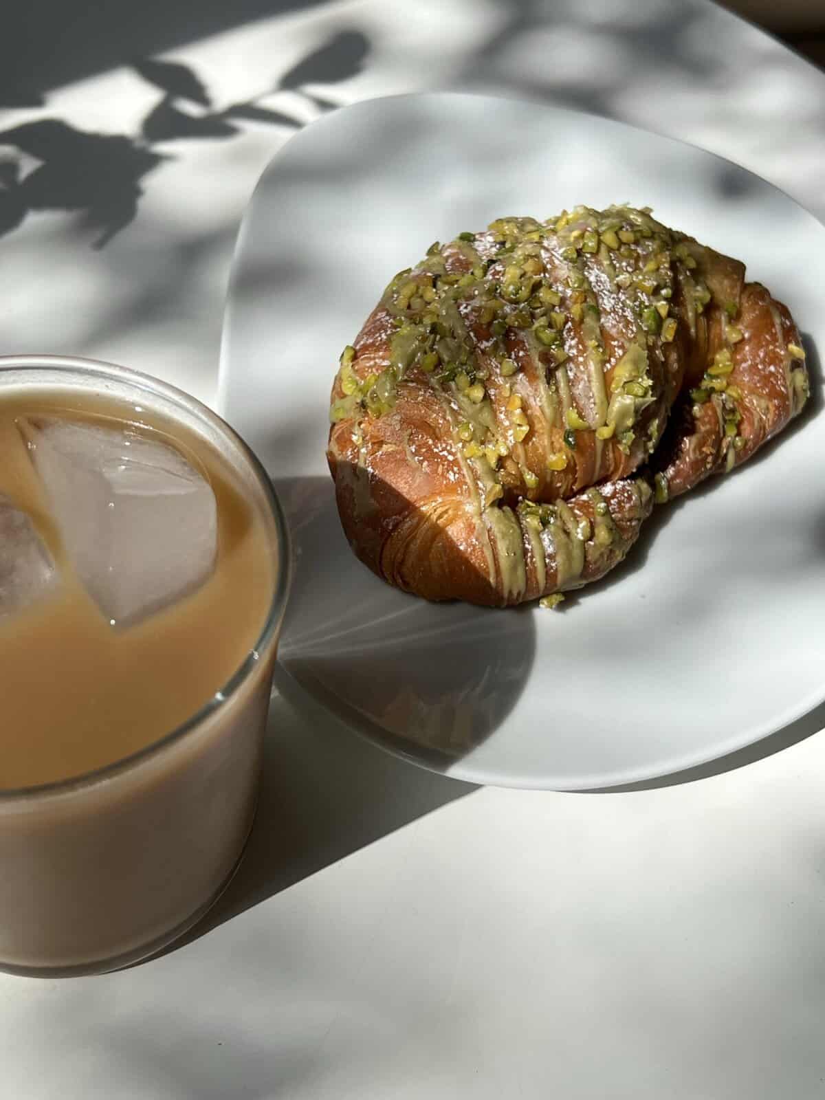Croissant topped with chopped pistachios on white plate, iced coffee drink on light background.