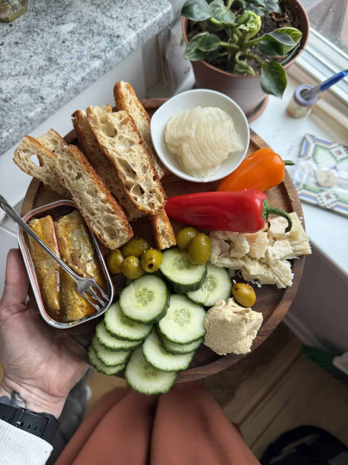 Fresh vegetable and bread platter with hummus, cheese, peppers, tinned fish and pickles, perfect for a healthy snack.